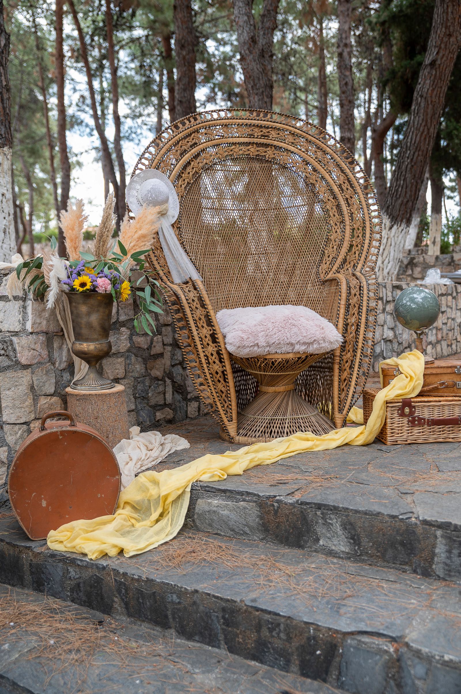 Wicker peacock chair with a pink cushion, a flower vase and yellow fabric on stone steps.