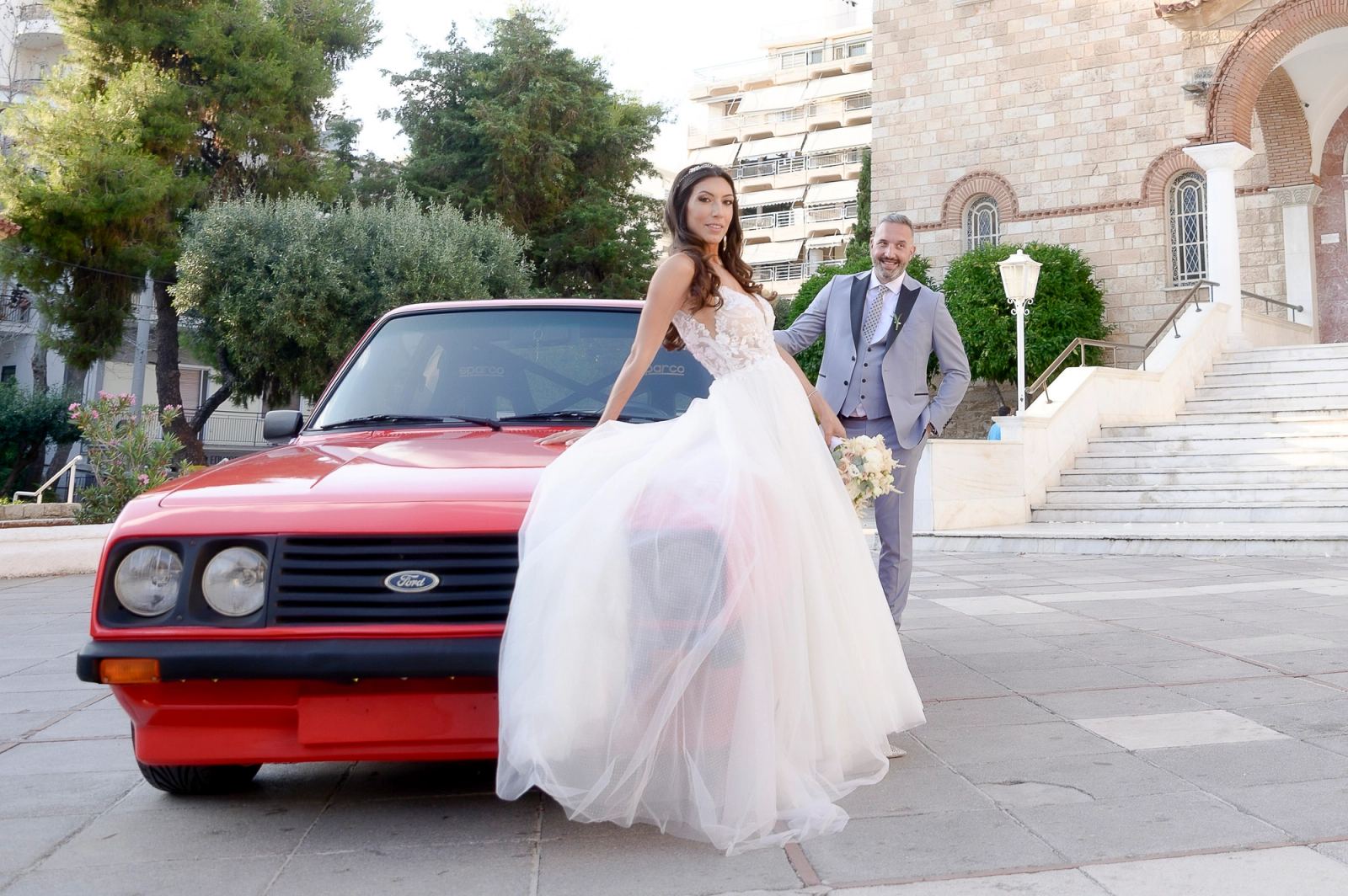 Η νύφη ακουμπά σε κόκκινο Ford μπροστά σε πέτρινη εκκλησία, ενώ ο γαμπρός με γκρι κοστούμι στέκεται πίσω της.