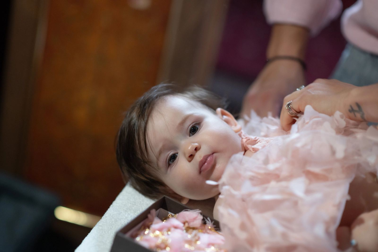 The baby in a pink feathered dress lies on a table as hands undress her.