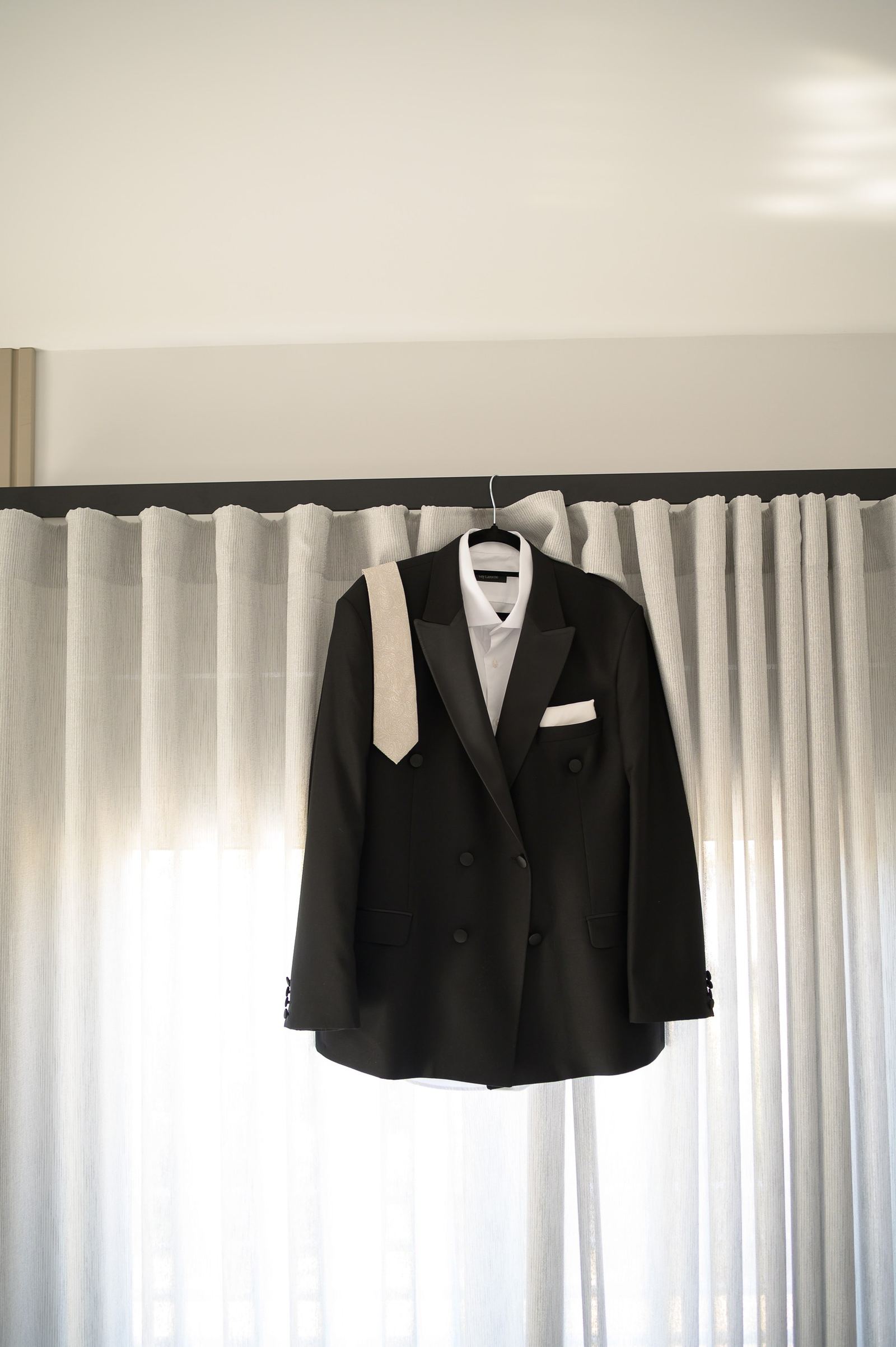 A black double-breasted jacket with a white shirt and silver tie hanging against a bright curtain.