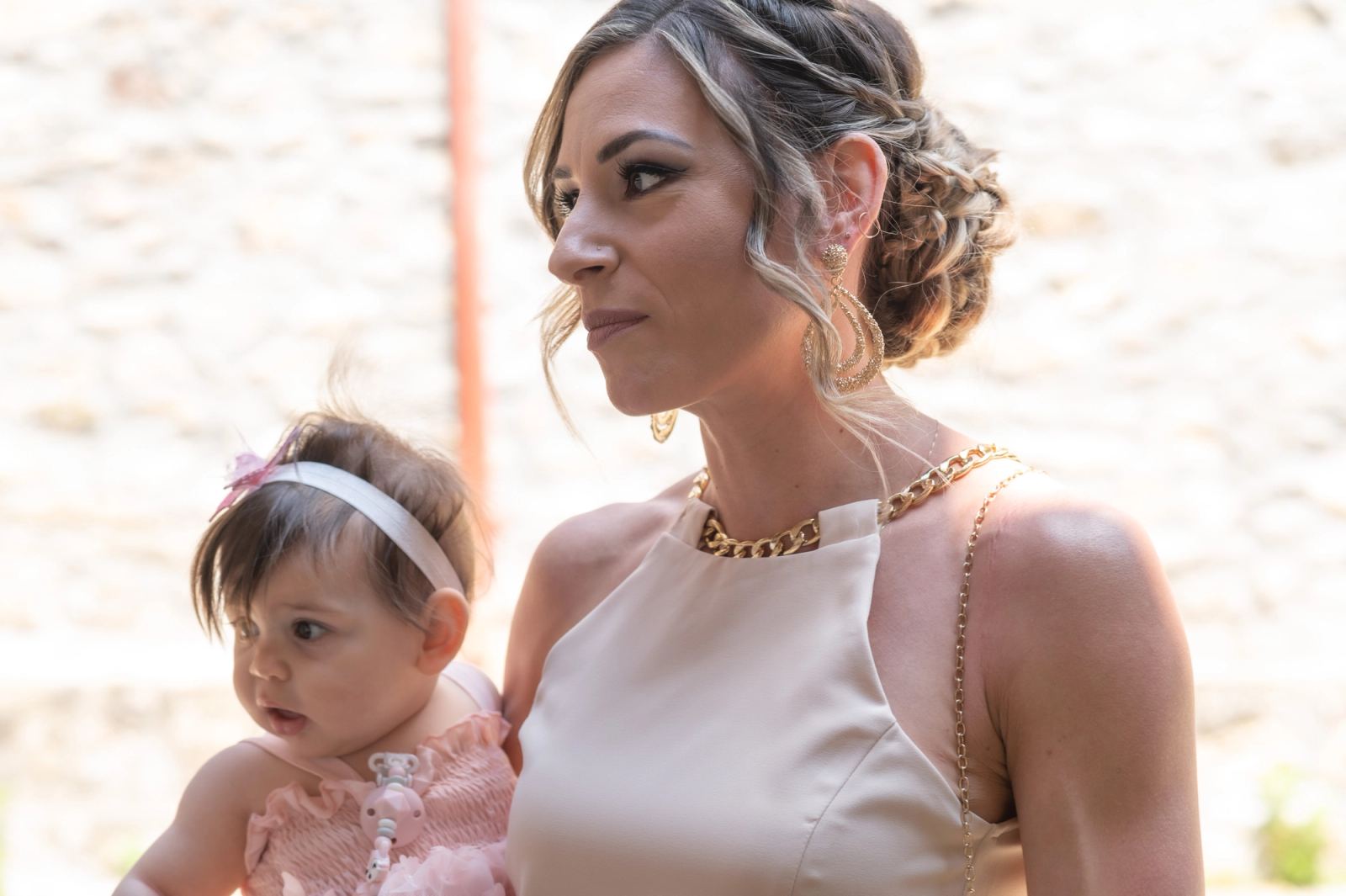A woman with a gold necklace and braided hair holds the baby in a pink dress and headband.
