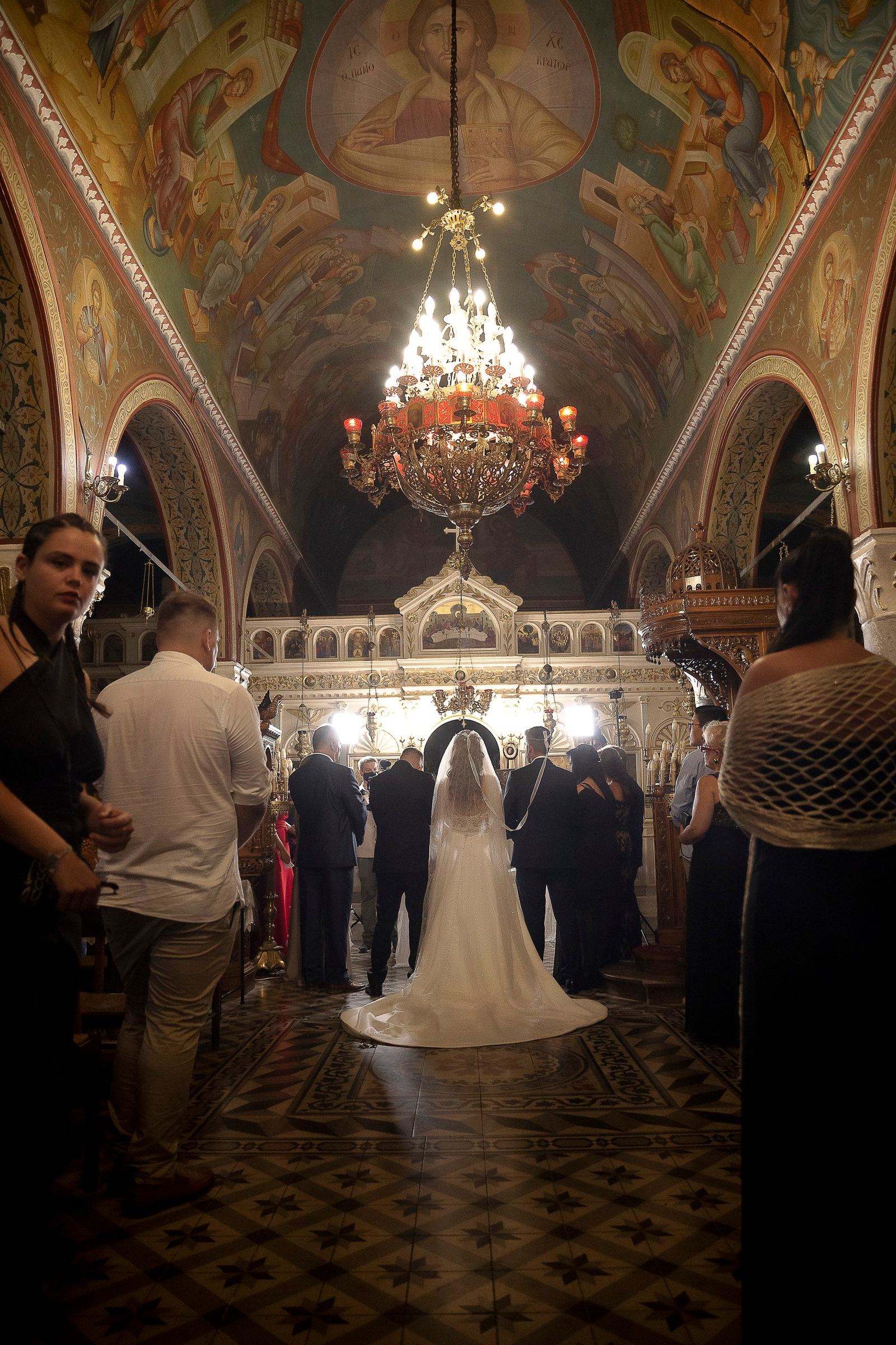 Η νύφη και ο γαμπρός μπροστά στο τέμπλο, κάτω από μεγάλο πολυέλαιο, με καλεσμένους στα πλάγια.