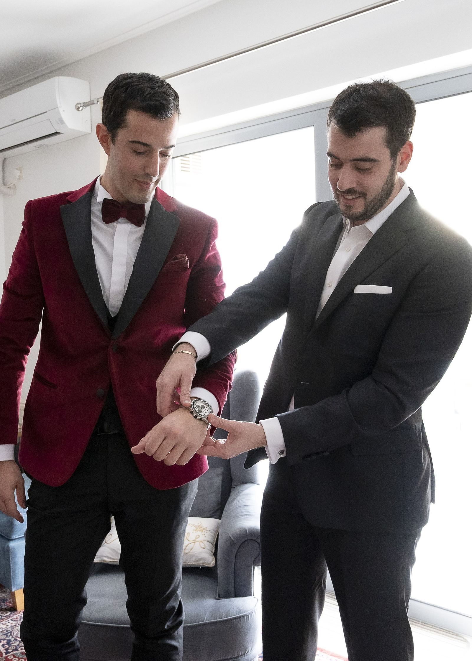 The groom in a burgundy velvet jacket as a man in a dark suit fastens his watch.