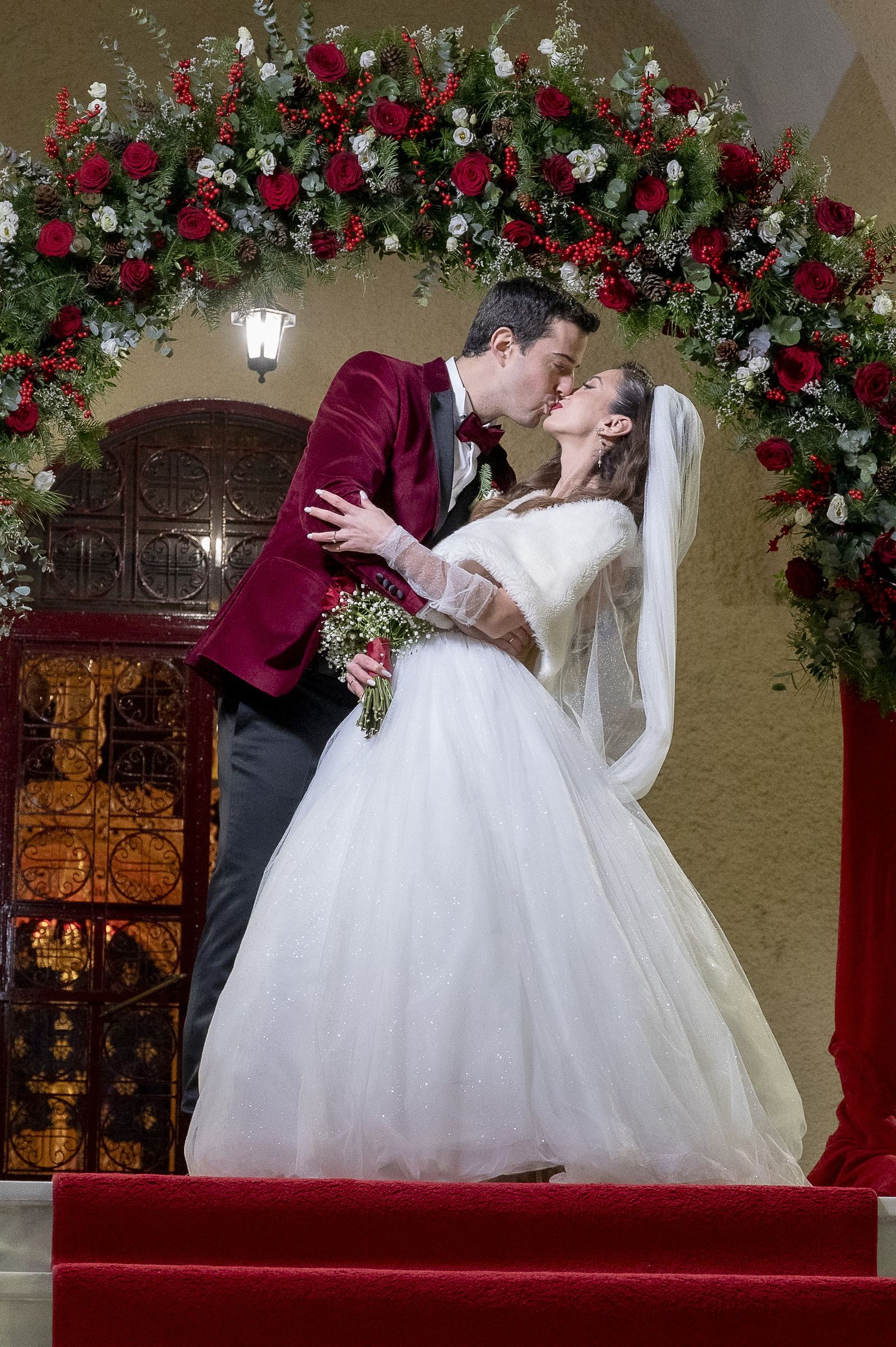The groom in a burgundy velvet jacket kisses the bride under an arch of red roses at a church entrance.