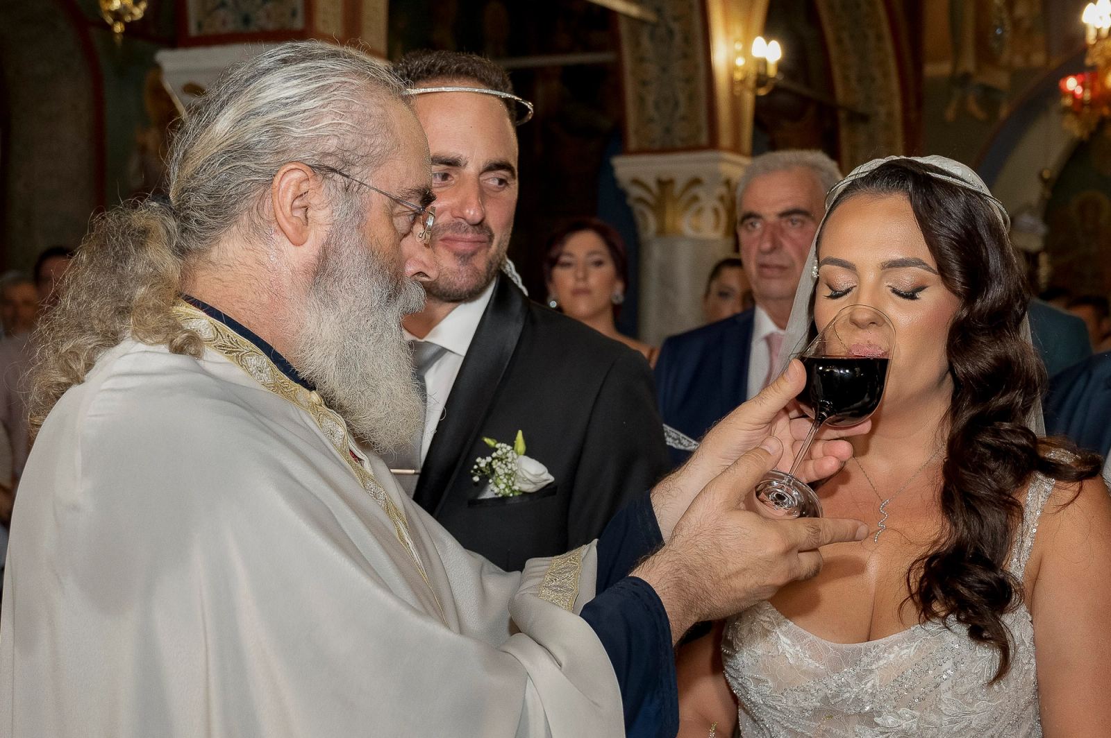 Ο ιερέας δίνει στη νύφη να πιει κόκκινο κρασί από ποτήρι, με τον γαμπρό δίπλα της.