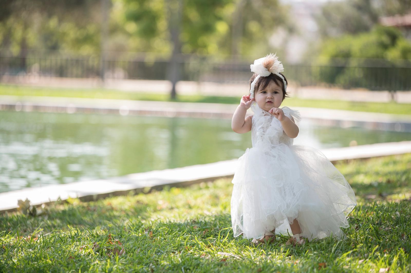 The baby in a white tulle dress stands on the grass beside a pond in a park.