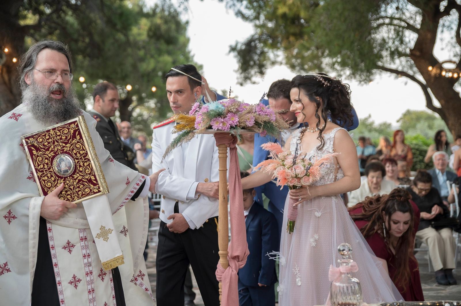Ο ιερέας με Ευαγγέλιο οδηγεί τη νύφη και τον γαμπρό γύρω από το τραπέζι, με λαμπάδα με ροζ λουλούδια.