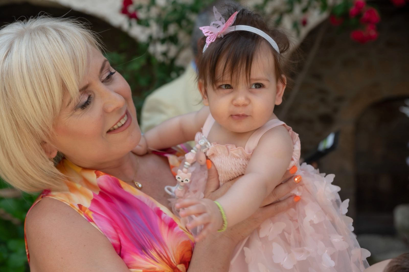 A blonde woman in a colorful dress holds the baby wearing a pink dress and a butterfly headband.
