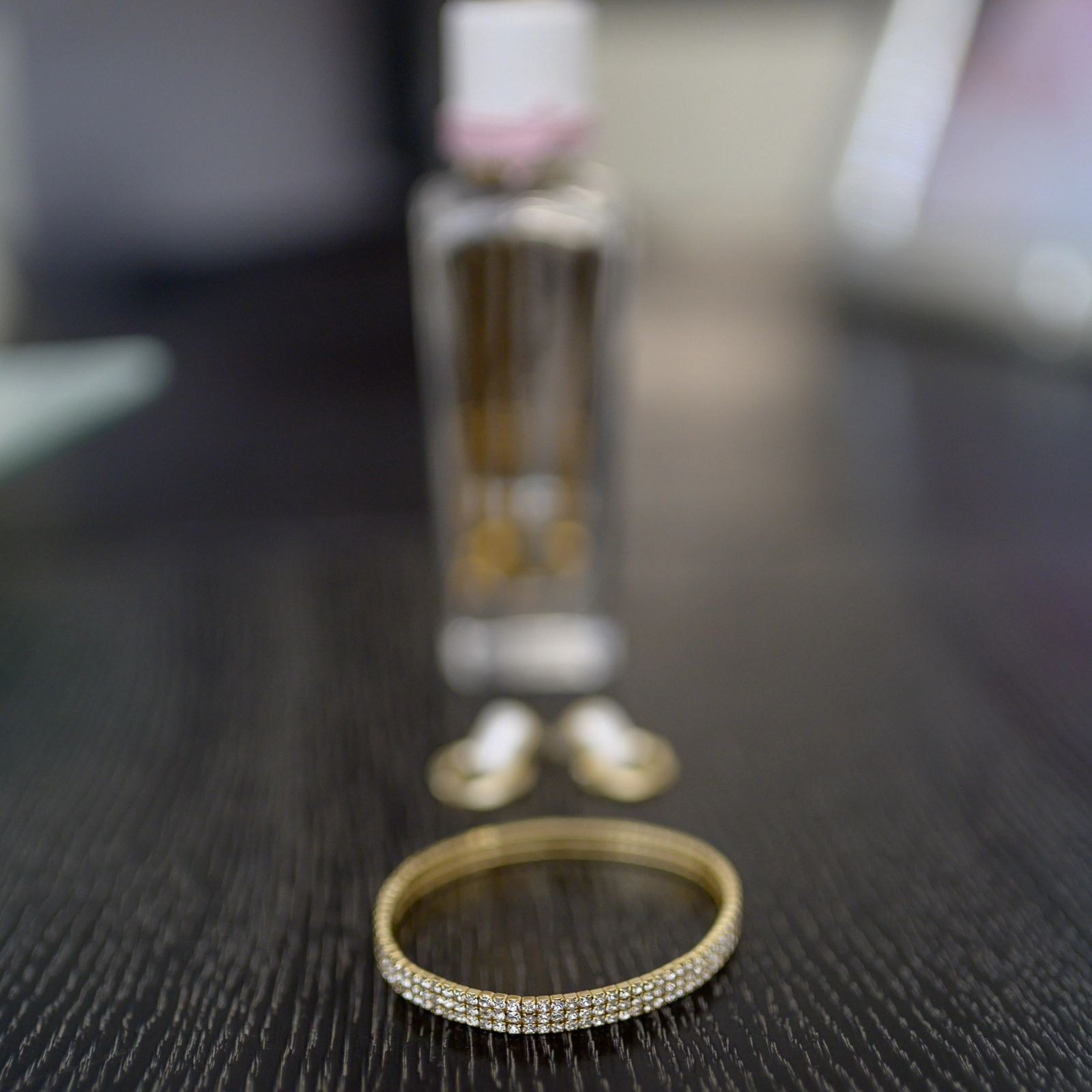A gold rhinestone bracelet and earrings on a dark wooden table, with a blurred perfume bottle behind.