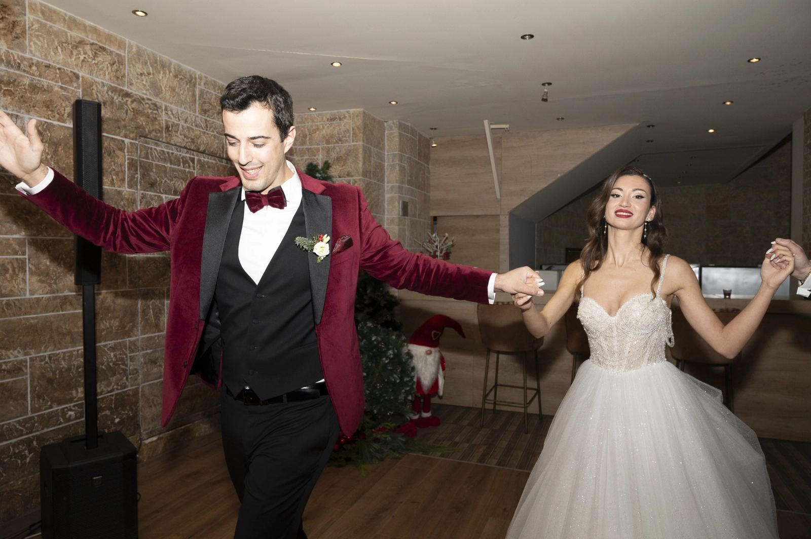 The groom and bride dance holding hands in a hall with stone walls and a wooden floor.