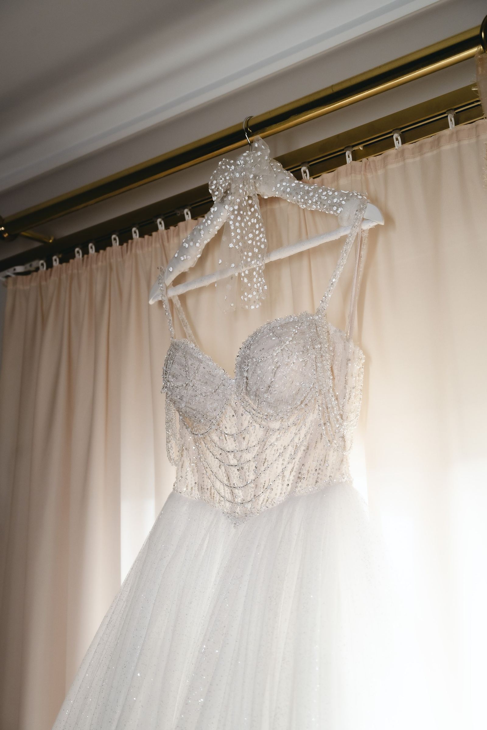 A wedding dress with a beaded bodice and tulle skirt hangs in front of pale curtains.