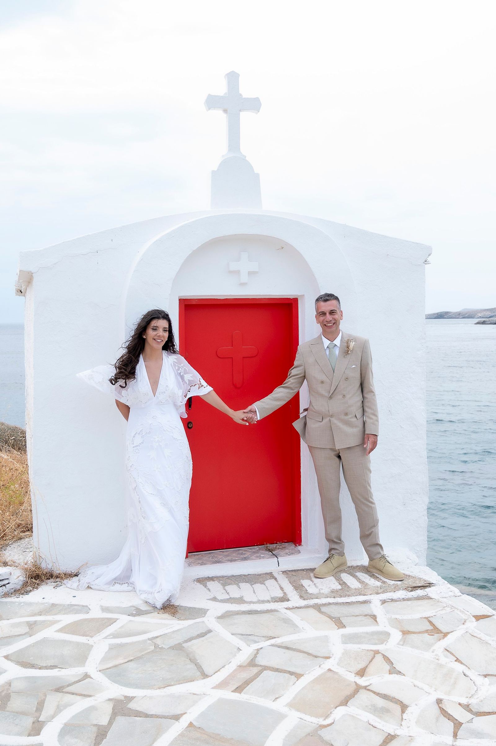 Η νύφη και ο γαμπρός κρατιούνται από το χέρι μπροστά στην κόκκινη πόρτα λευκού εκκλησακιού δίπλα στη θάλασσα.