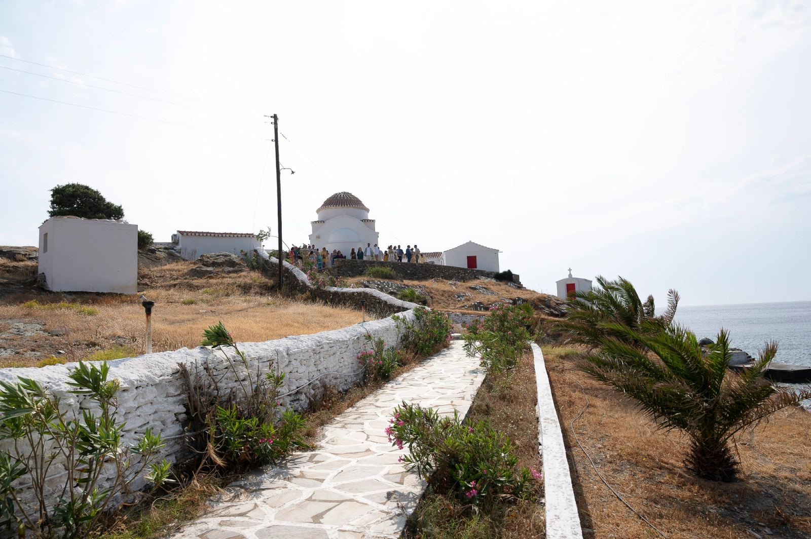 Πλακόστρωτο μονοπάτι με λευκό τοιχάκι οδηγεί σε λευκή εκκλησία με τρούλο δίπλα στη θάλασσα.