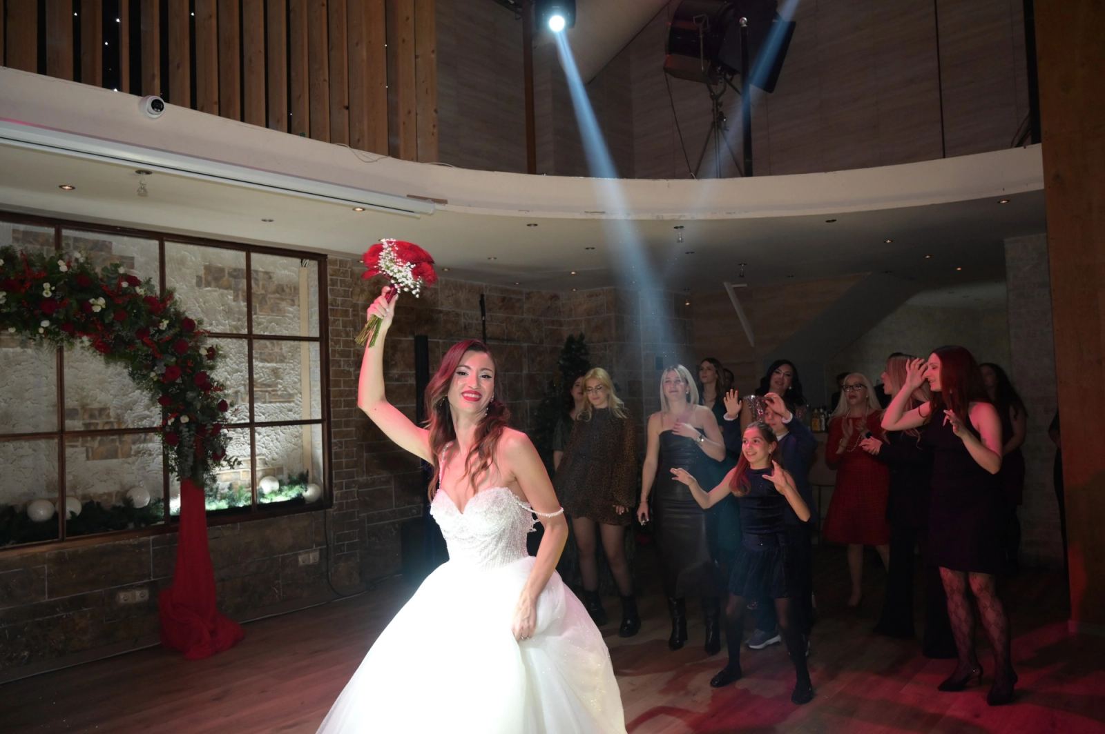 The bride raises a bouquet of red roses while female guests wait behind her on the dance floor.