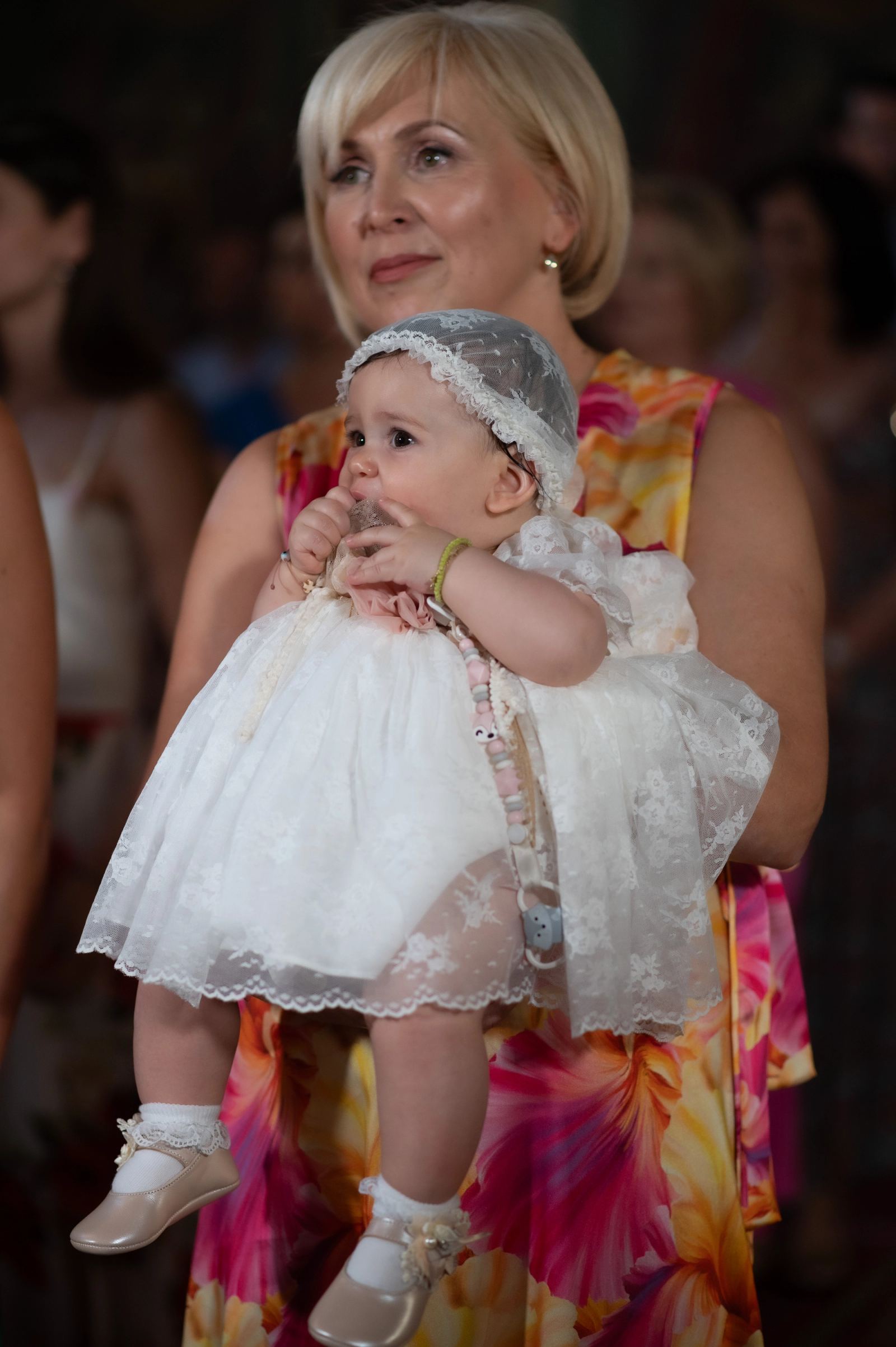 A blonde woman in a floral dress holds the baby in a white lace dress and bonnet.