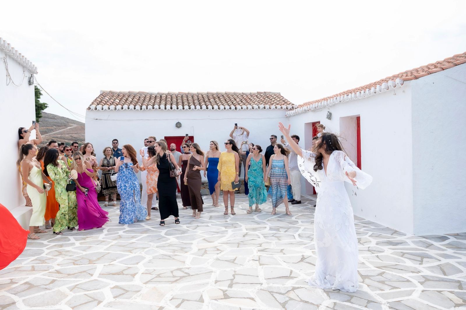Η νύφη με τα χέρια ψηλά γυρίζει προς τις καλεσμένες, που κοιτούν ψηλά σε λευκή αυλή με κεραμοσκεπές.