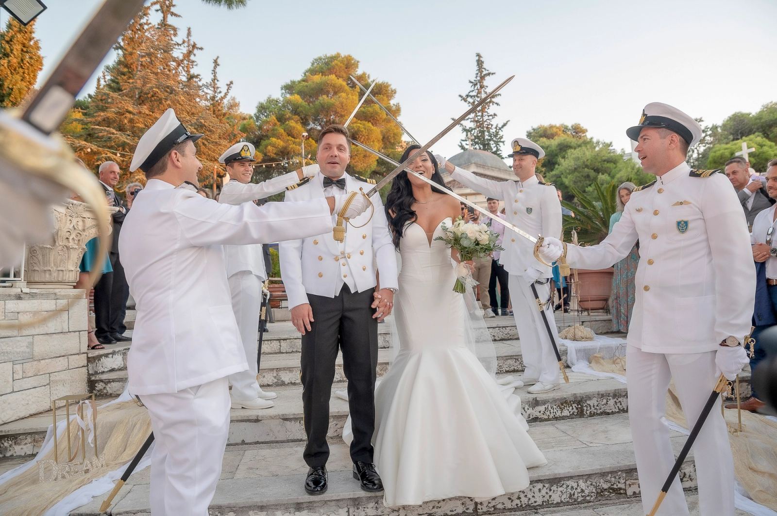 Η νύφη και ο γαμπρός στέκονται κάτω από σταυρωμένα σπαθιά ναυτικών σε πέτρινα σκαλιά με δέντρα πίσω.