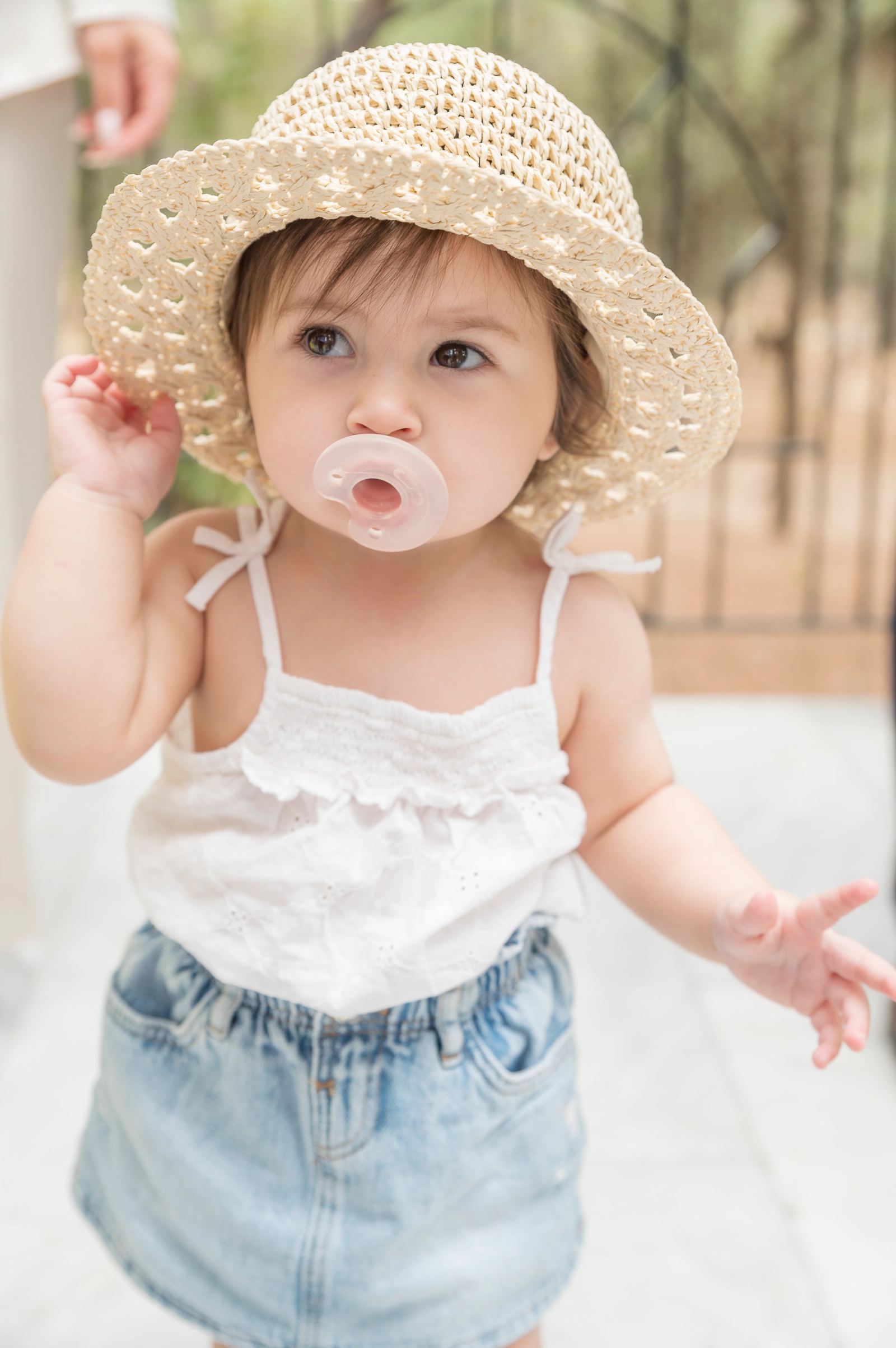 The baby in a straw hat, white top, denim skirt and pacifier stands in a courtyard.