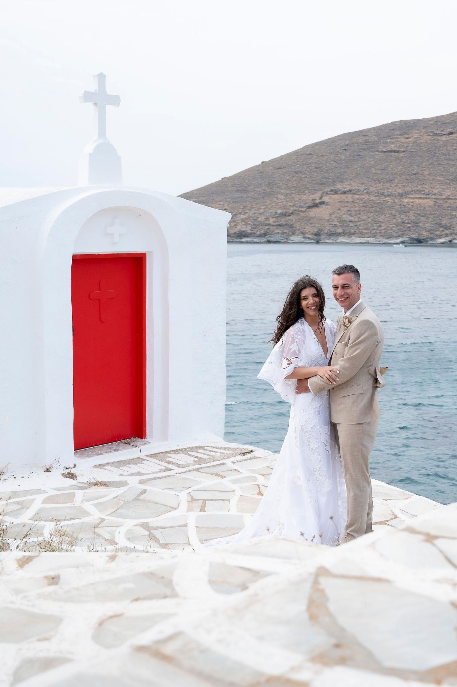 Η νύφη και ο γαμπρός αγκαλιασμένοι δίπλα σε λευκό εκκλησάκι με κόκκινη πόρτα, με θάλασσα και λόφο πίσω.