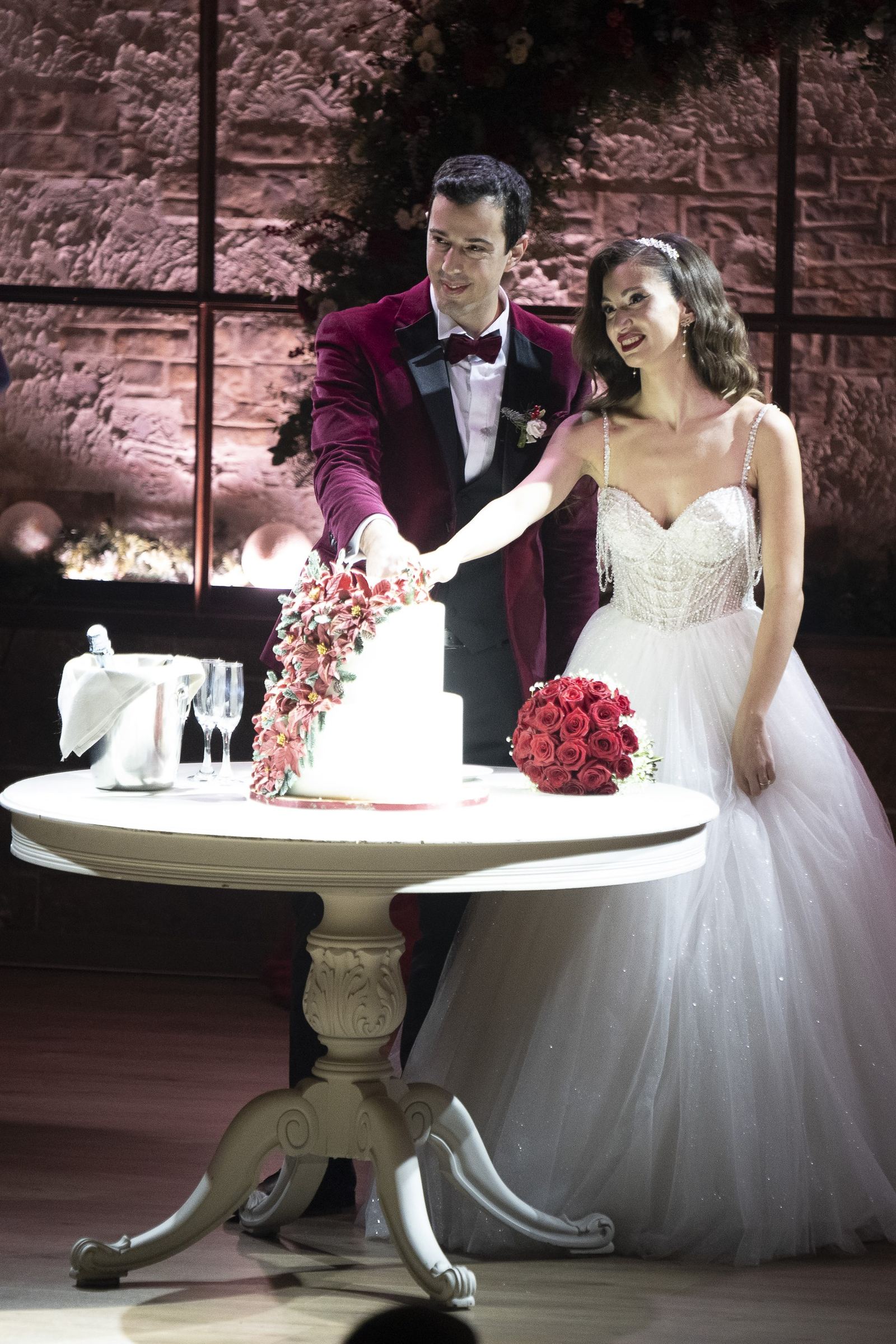 The bride and groom cut a white cake with red flowers on a round white table.