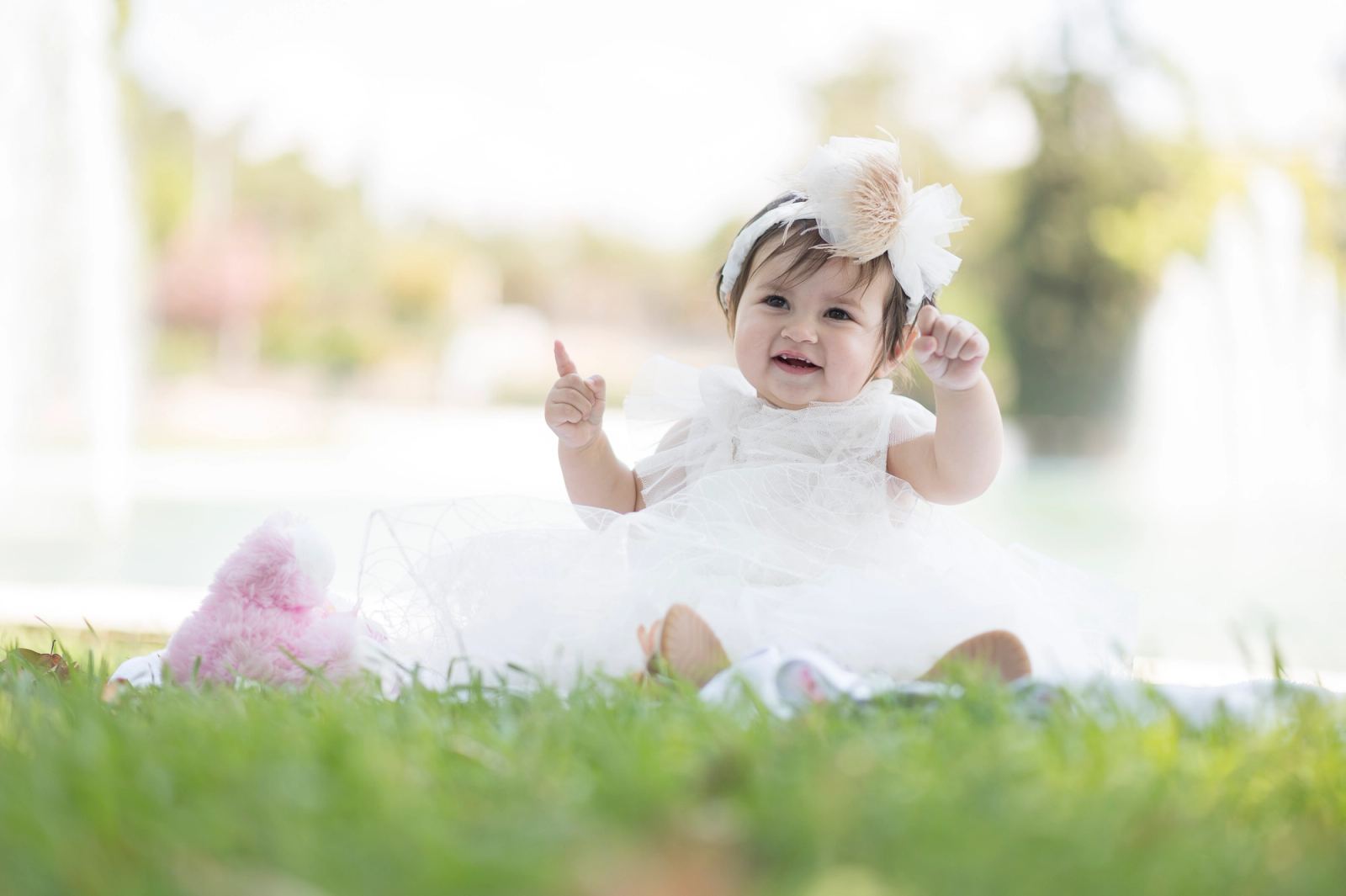 The baby in a white dress and feathered headband sits smiling on the grass, a fountain behind.