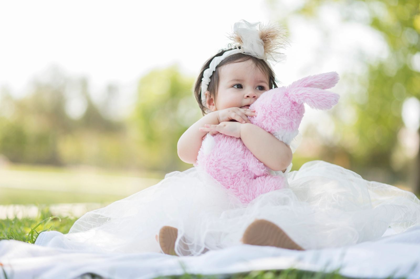 The baby in a white tulle dress sits on the grass hugging a pink plush bunny.
