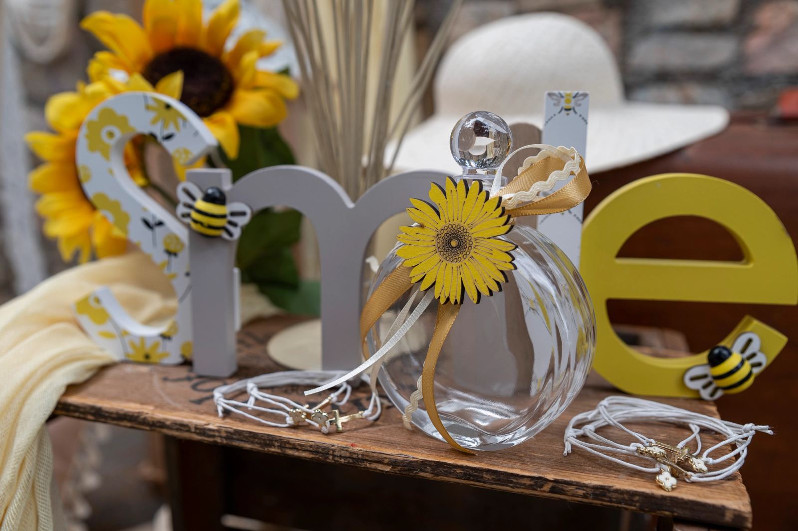 Glass bottle with a yellow daisy, witness bracelets and wooden letters with little bees on a wooden table.