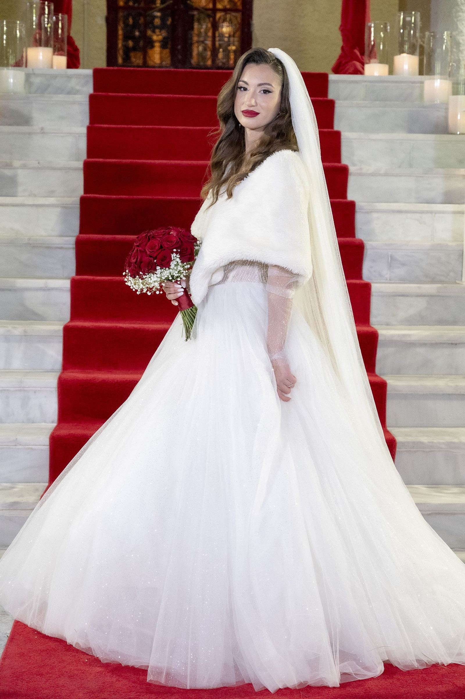 The bride in a white tulle gown holding red roses stands on marble steps with a red carpet.