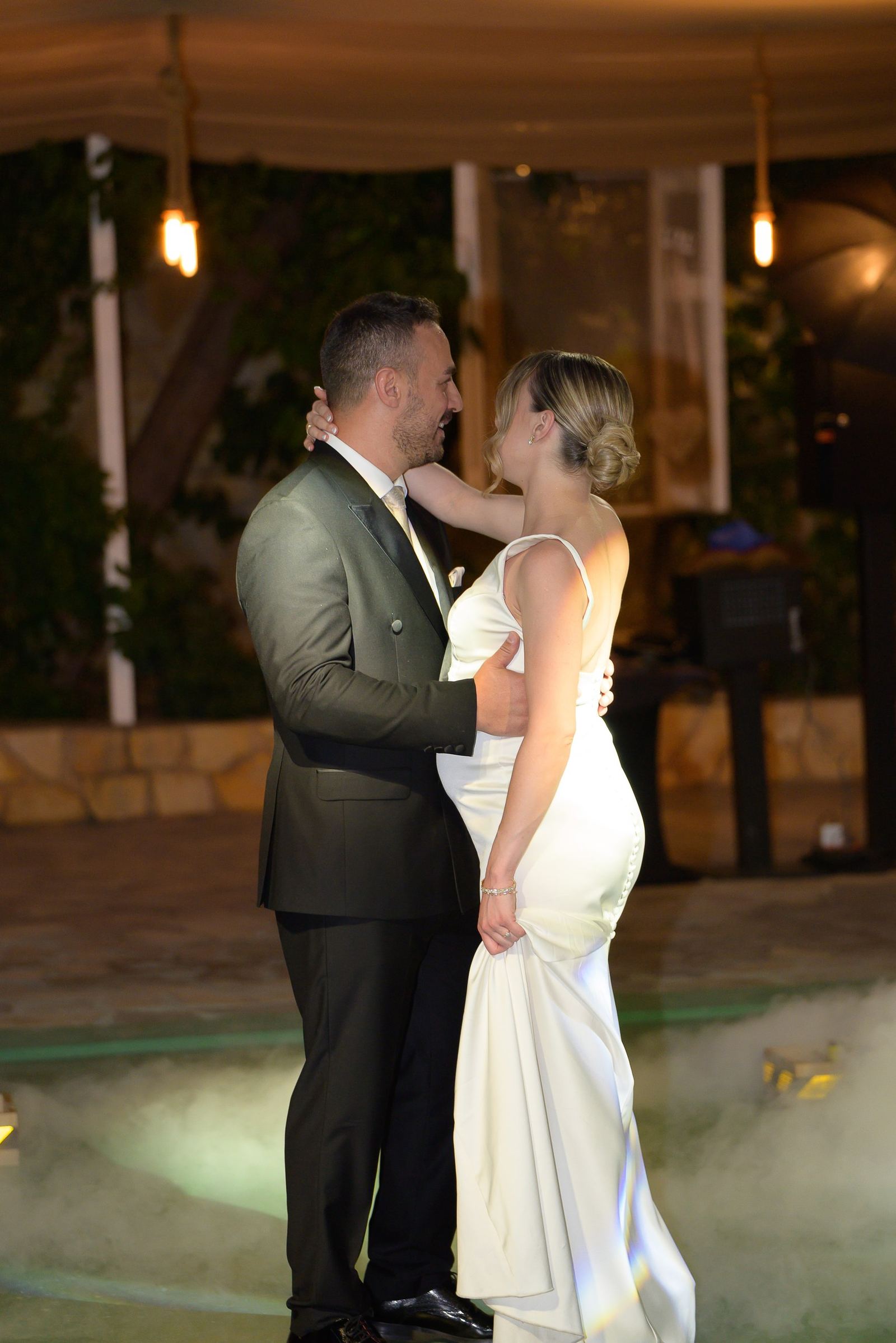 The bride in a satin gown and the groom dance in an embrace above low fog on the dance floor.