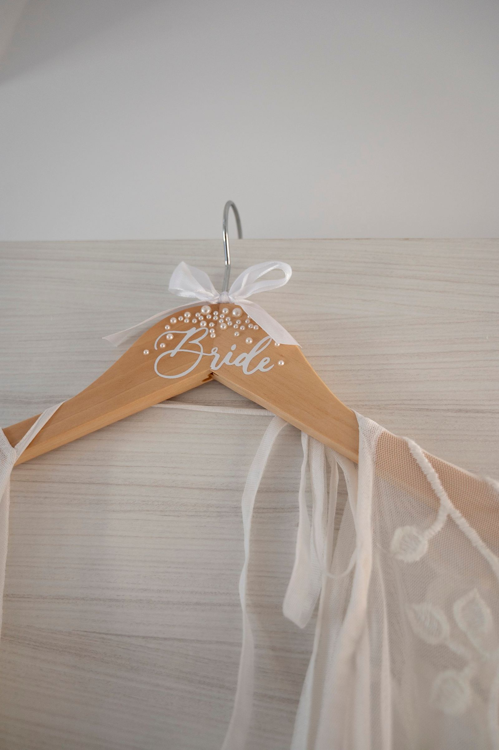 Ξύλινη κρεμάστρα με τη λέξη «Bride», πέρλες και άσπρη κορδέλα, με δαντελένιο νυφικό κρεμασμένο.