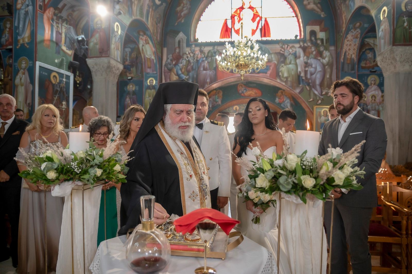 Ο ιερέας διαβάζει μπροστά στο τραπέζι του μυστηρίου, με το ζευγάρι πίσω του σε αγιογραφημένη εκκλησία.