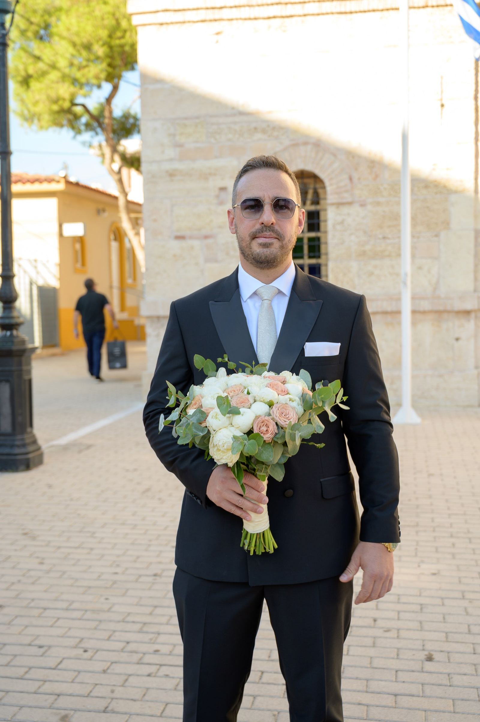 The groom in a black suit and sunglasses holds a bridal bouquet in front of a stone church.