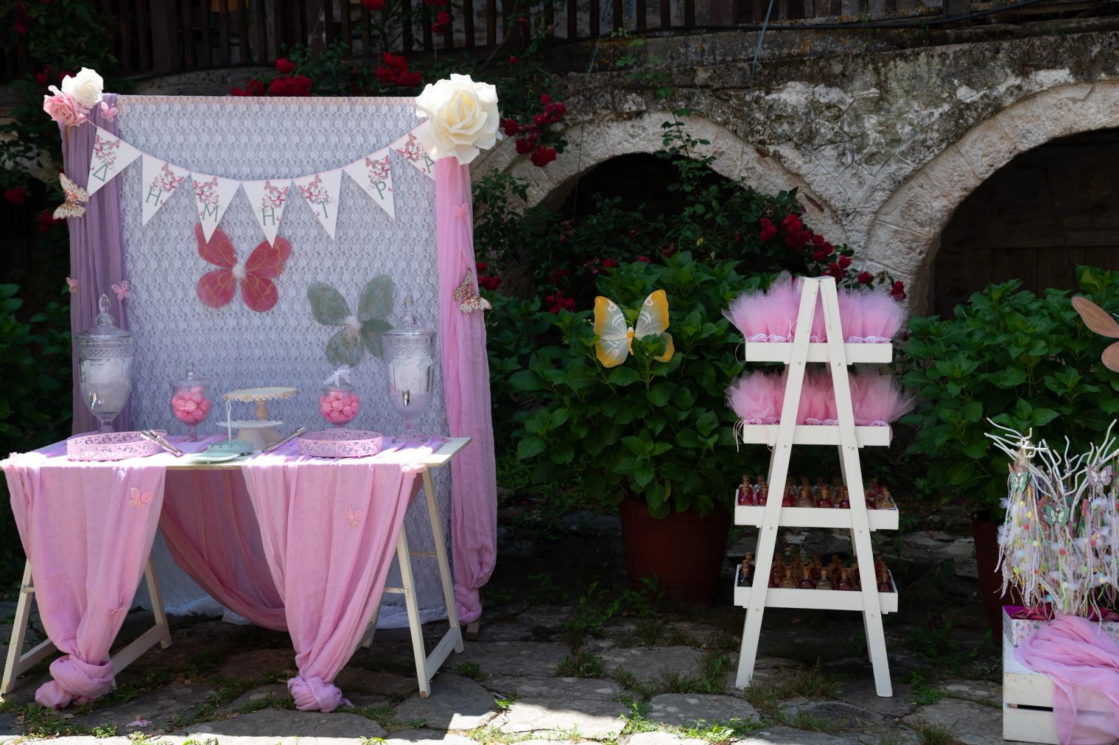 A table with pink drapes, sweets and butterflies beside a white ladder of favors in a stone courtyard.
