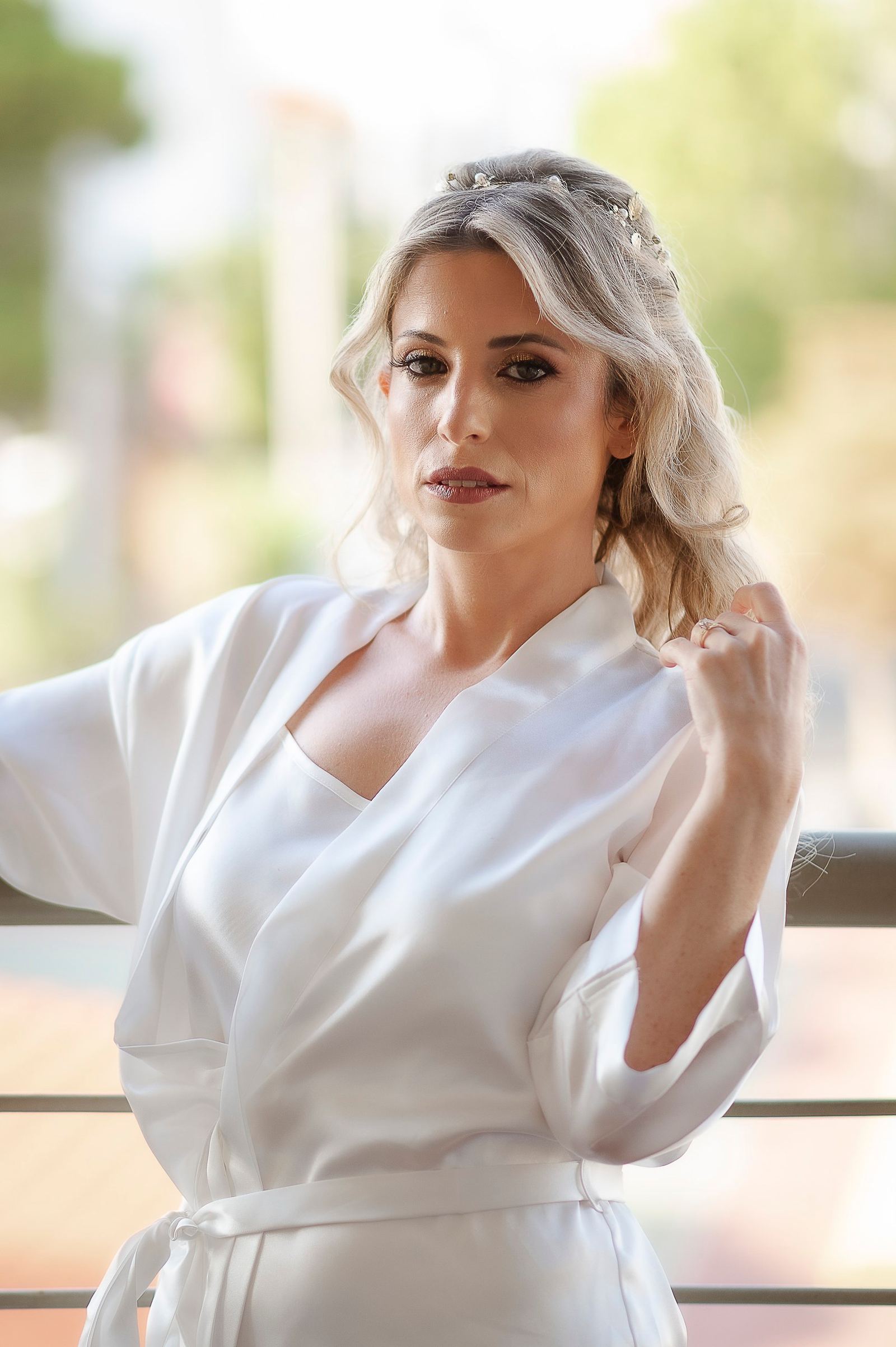 Η νύφη με λευκή σατέν ρόμπα και στέκα στα μαλλιά ποζάρει σε μπαλκόνι με φωτεινό φόντο.