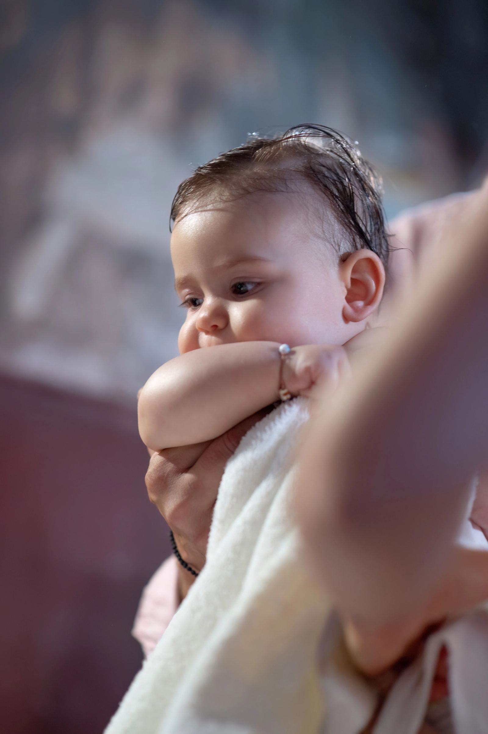 The baby with wet hair and a small bracelet, wrapped in a white towel, held by hands inside a church.