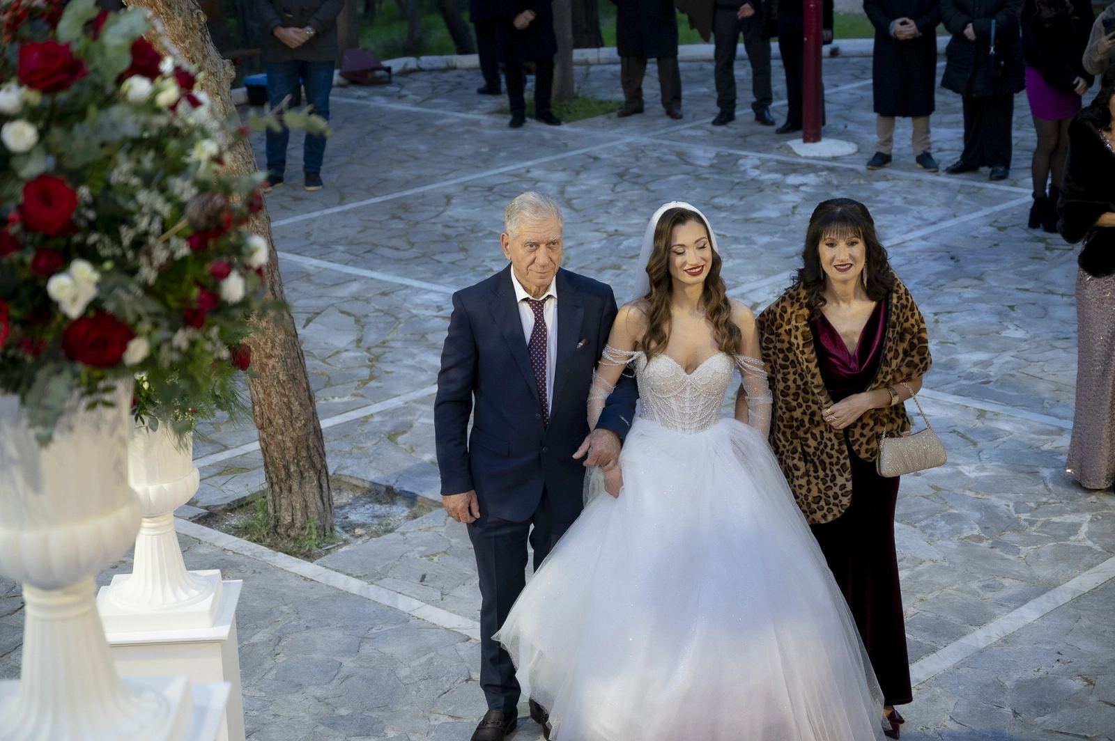 The bride walks with an older man and a woman in a leopard wrap through a courtyard with guests