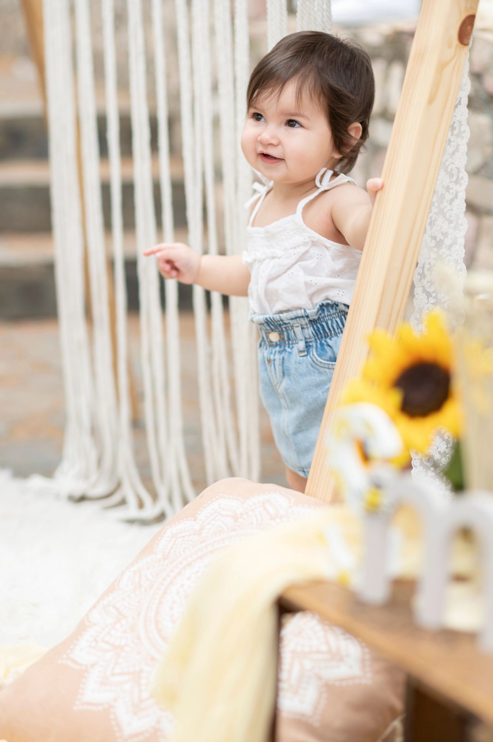 The baby stands by a wooden post pointing with one hand, a sunflower in front.
