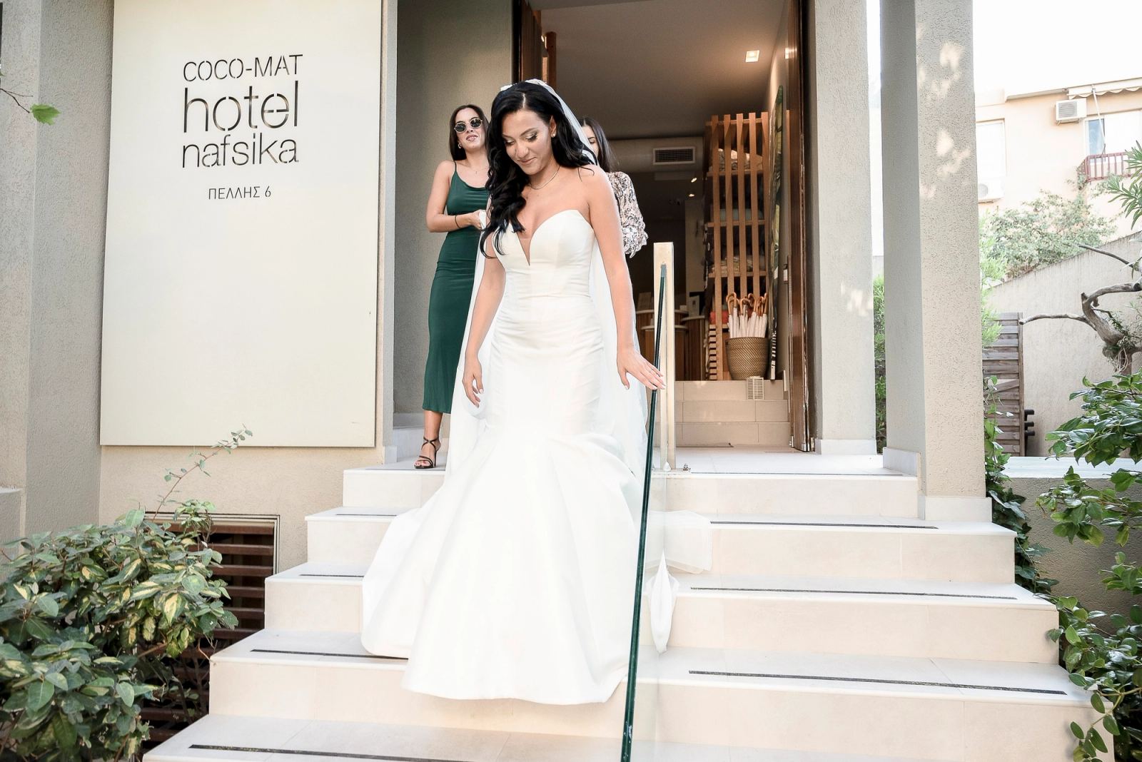Η νύφη κατεβαίνει τα σκαλιά του Coco-Mat Hotel Nafsika με δύο γυναίκες πίσω της.