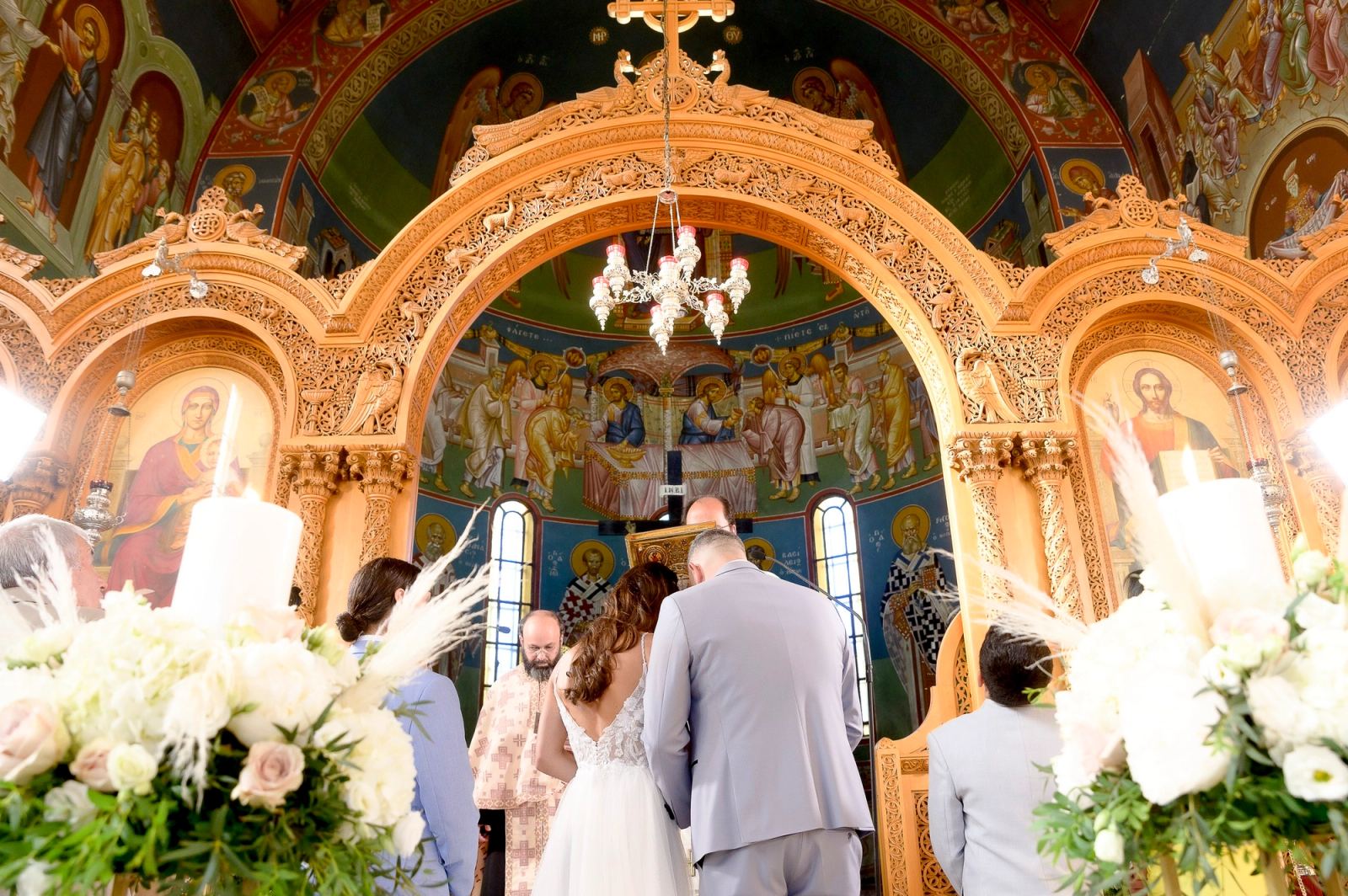 Η νύφη και ο γαμπρός από πίσω μπροστά σε σκαλιστό επίχρυσο τέμπλο με ψηφιδωτά και λαμπάδες.