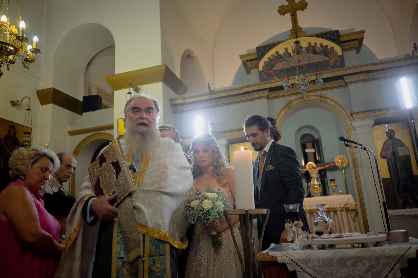 Ο ιερέας με Ευαγγέλιο στέκεται με τη νύφη και τον κουμπάρο μπροστά στο τέμπλο μιας φωτεινής εκκλησίας.