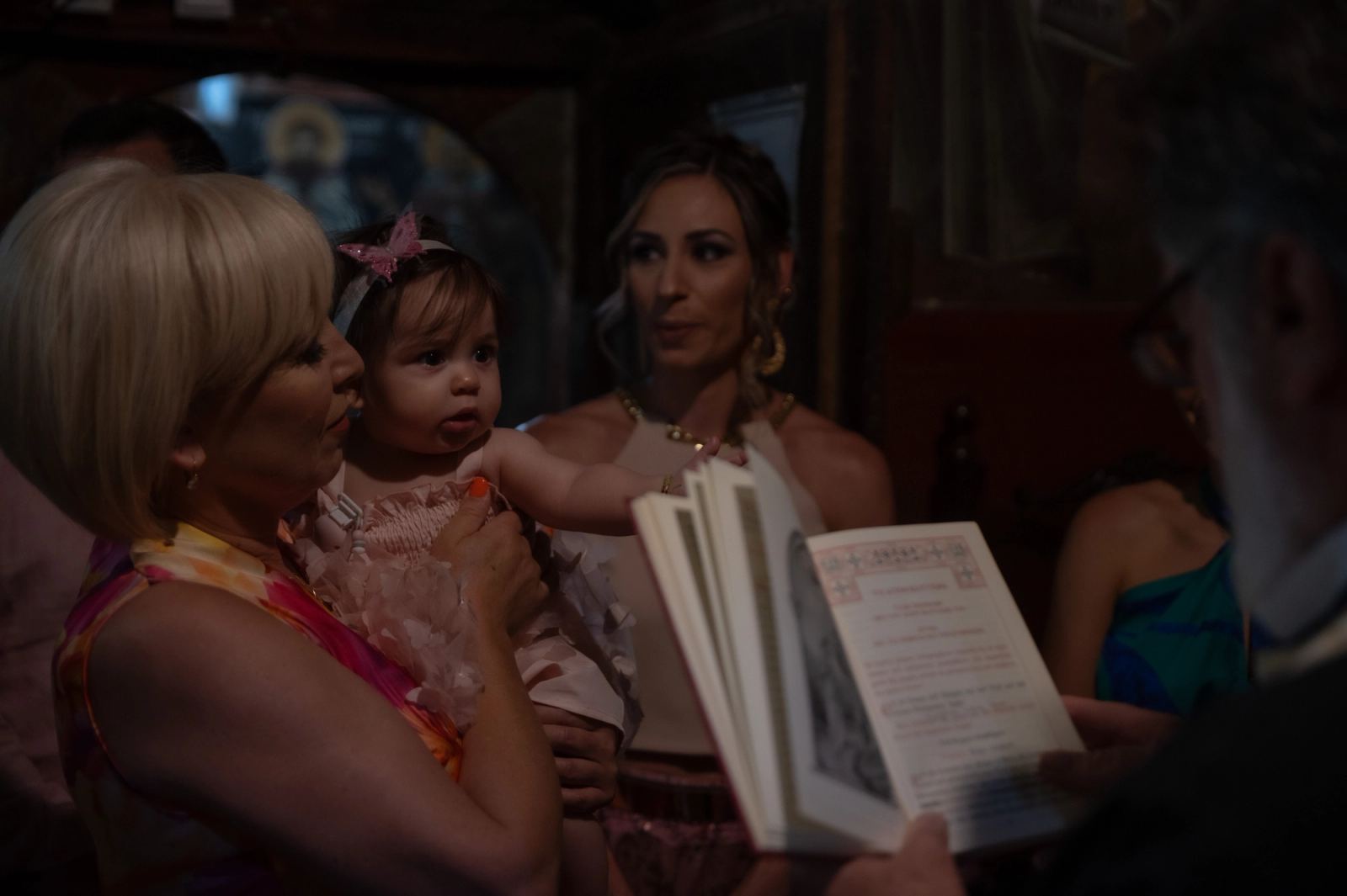 A woman holds the baby in the dim church as the priest reads from an open book.