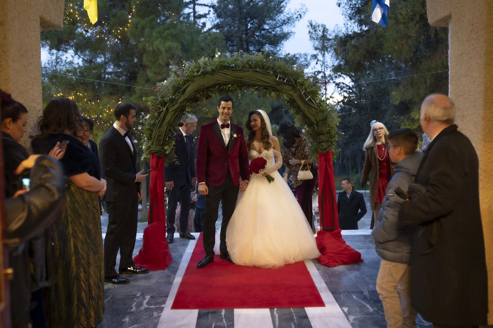 The bride and groom under a greenery arch on a red carpet, surrounded by guests