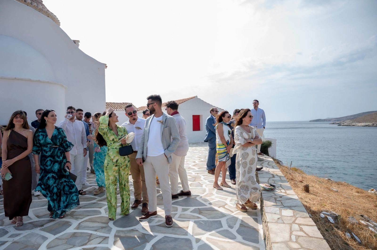 Καλεσμένοι με καλοκαιρινά ρούχα σε πλακόστρωτη αυλή λευκής εκκλησίας δίπλα στη θάλασσα.
