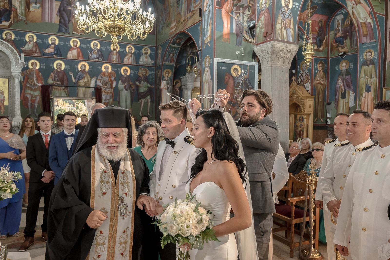 Ο ιερέας κρατά το χέρι του γαμπρού ενώ ένας άντρας με γκρι κοστούμι αλλάζει τα στέφανα σε εκκλησία με τοιχογραφίες.
