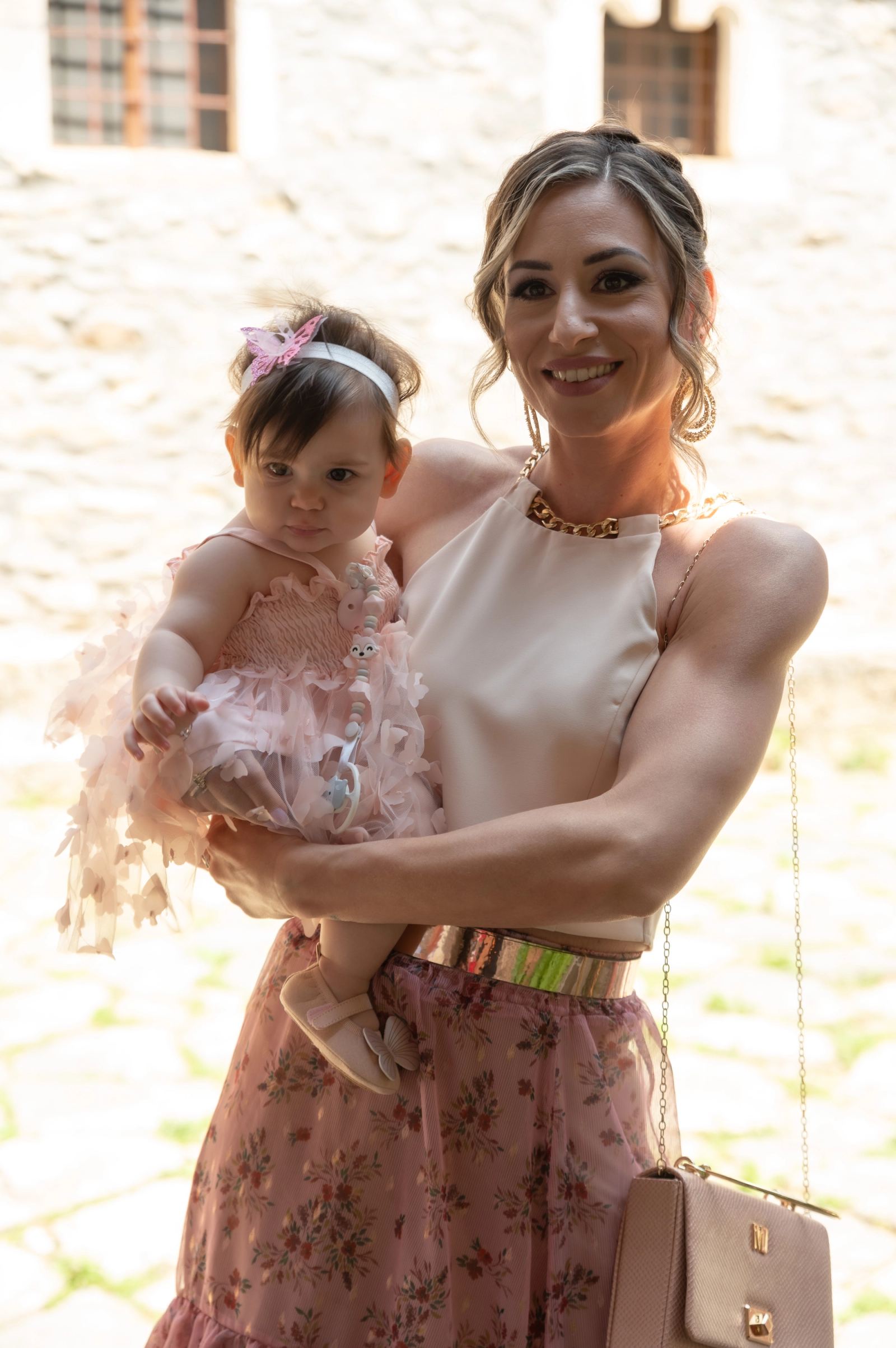 A woman holds the baby in a pink dress and headband in front of a stone wall.
