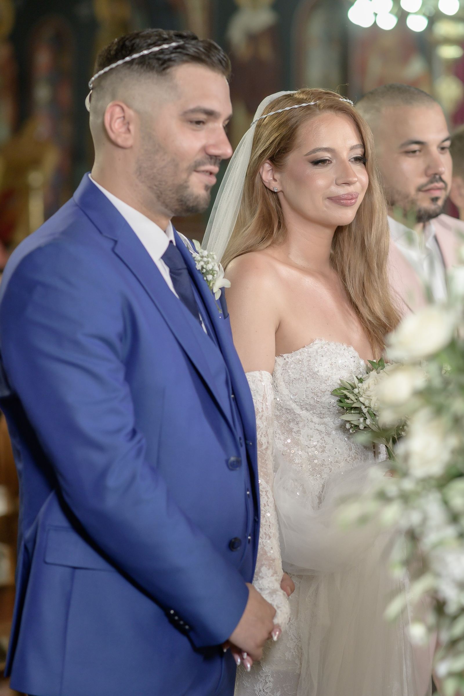 Ο γαμπρός και η νύφη με στέφανα στέκονται δίπλα δίπλα στην εκκλησία, με έναν καλεσμένο πίσω τους.