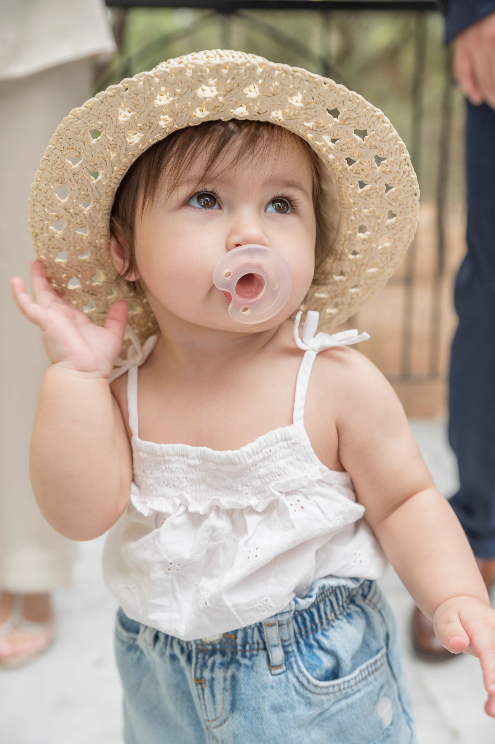 The baby in a straw hat, white top and pacifier raises a hand to the hat.