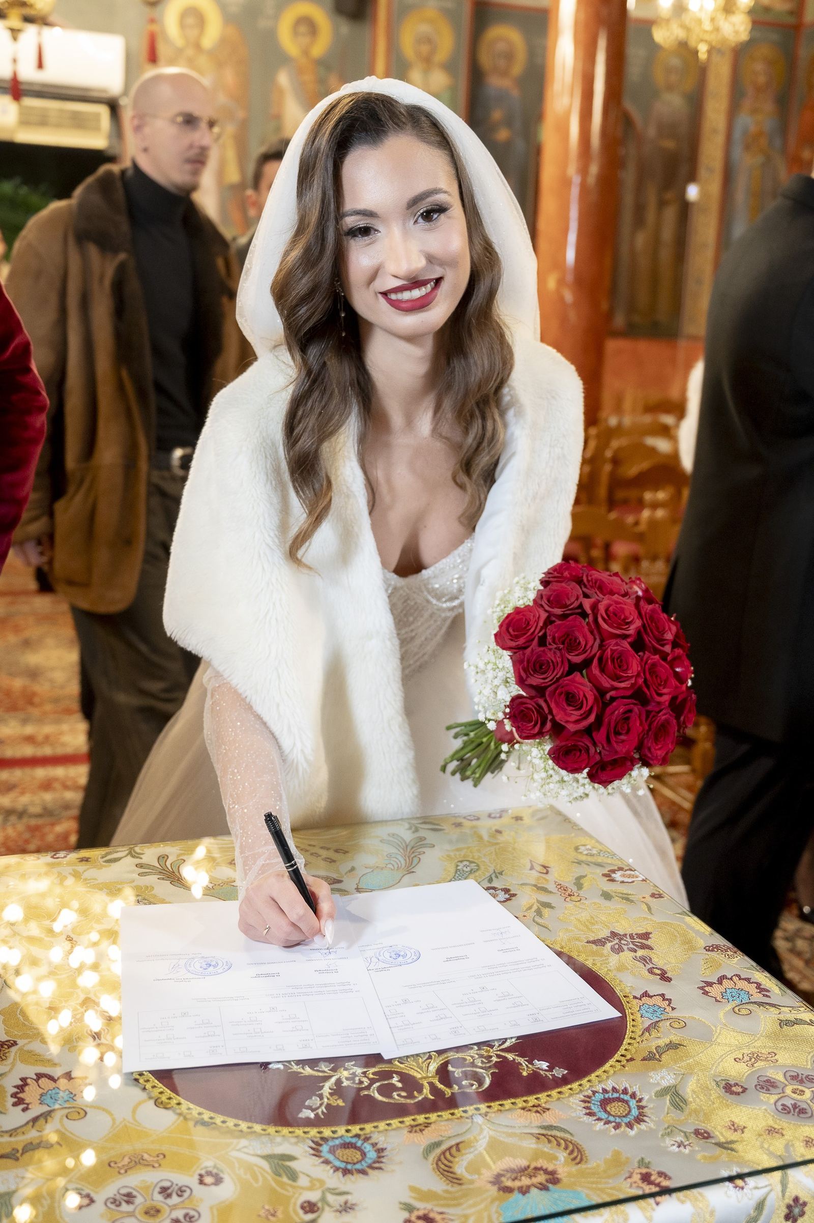 The bride in a white fur cape signs documents on a table with embroidered cloth inside a church with icons.