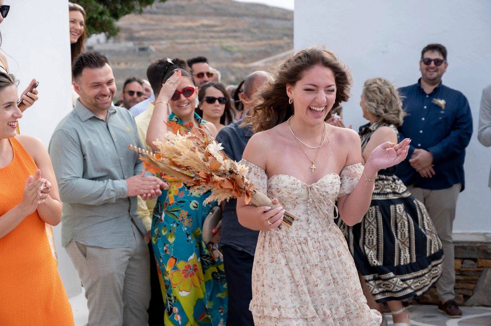 Μια καλεσμένη με φλοράλ φόρεμα γελά κρατώντας την ανθοδέσμη, ενώ γύρω της άλλοι καλεσμένοι χειροκροτούν.