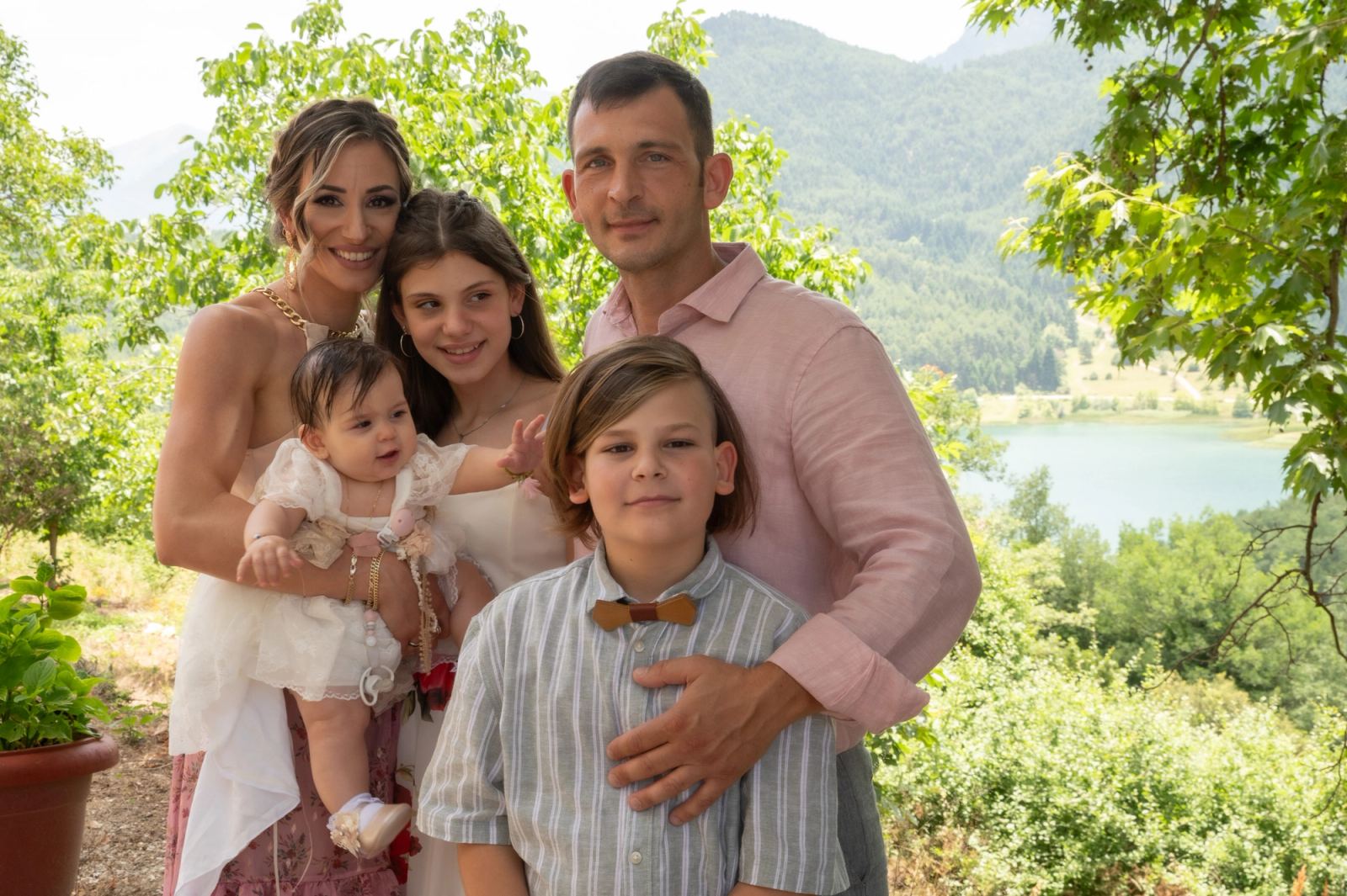 A family with the baby, a girl and a boy in a bow tie poses under trees overlooking a lake.
